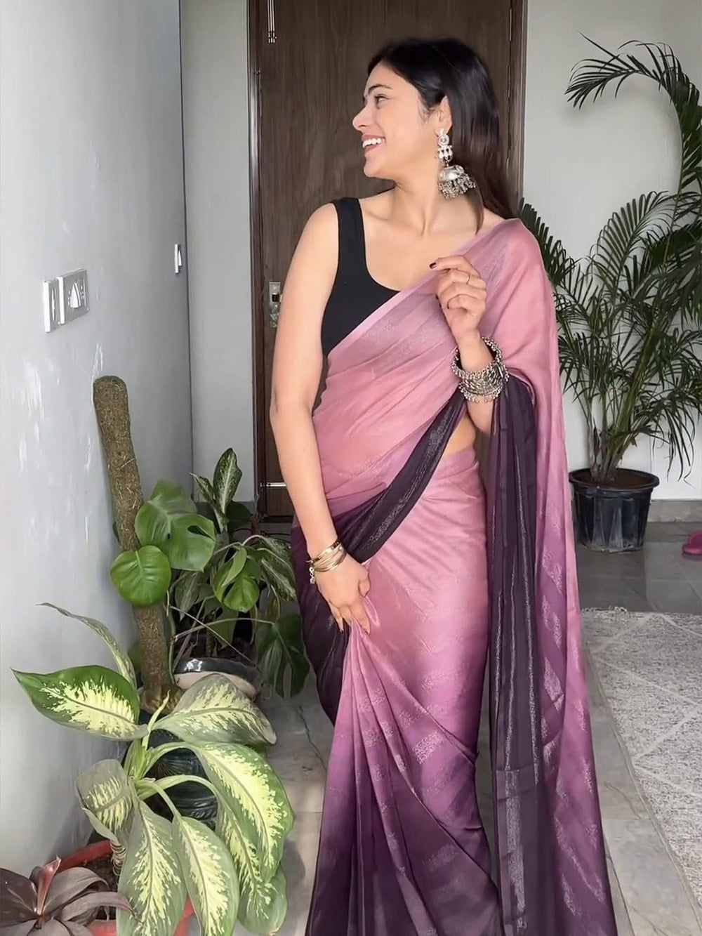 Woman in a pink saree with a black blouse standing indoors with plants around.