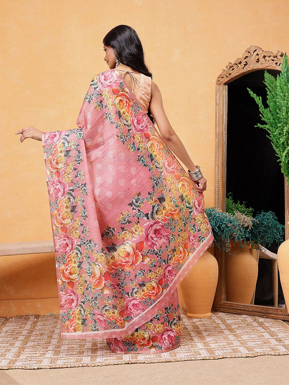 Woman wearing a pink floral saree in a room with a mirror and plants.