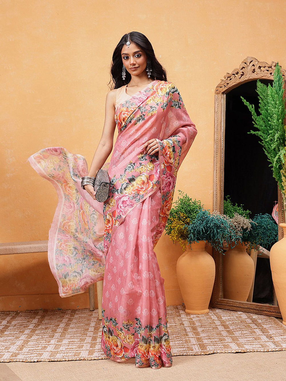 Woman in a pink saree with floral patterns standing in a room with decorative elements.